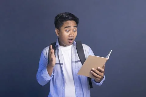 Shocked Asian student with backpack looking at an open notebook with mouth .. Stock Photos