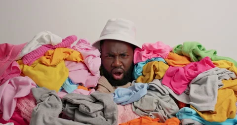 Shocked dark skinned bearded man in mess of laundry sticks head through dirty Stock Footage 156226843