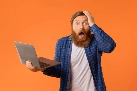 Shocked redhaired bearded man posing with laptop touches head, studio Foto stock