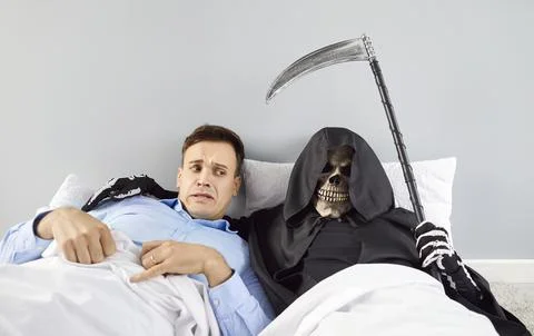 Shocked scared man lying on bed with skeleton in a grim reaper death costume Stockfoto's