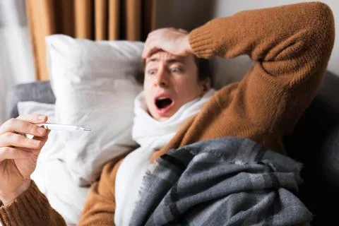 Shocked sick man lying on sofa holding thermometer and reacting to high fev.. Stock Photos
