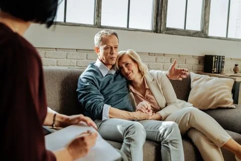 Short-haired old man supporting his crying wife while explaining problem Stock Photos
