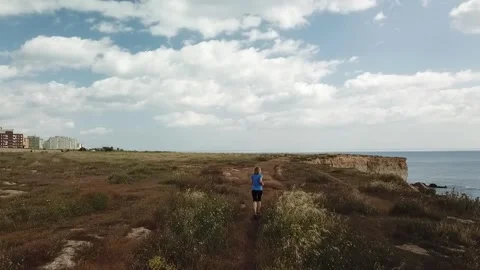 The shot captures the runner moving past dry grass and rocky edges by the sea Stock Footage 346449550