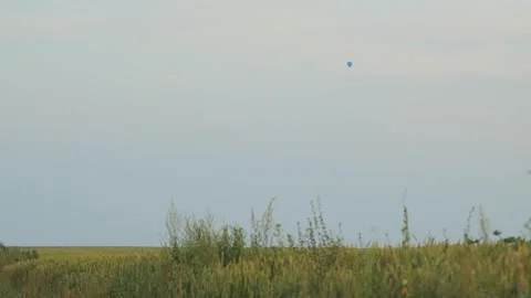 Shot during sunset, a blue balloon flies across the sky over a green field Stock Footage 247060124