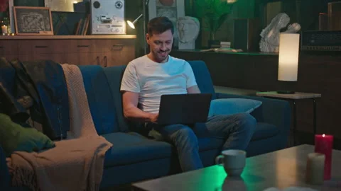 Shot of smiling handsome man using at laptop, chatting while sitting on sofa in Stock Footage 232872474