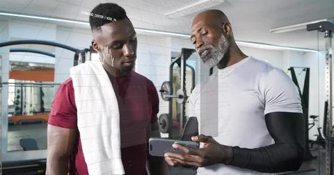 Showing trainer guiding trainee in weight room in sportswear with smartphone, 스톡 사진