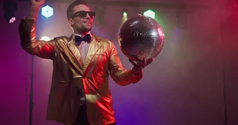 Showman in gold jacket holding disco ball under colorful stage lights at party Stock Footage 329289876
