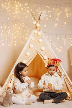 Siblings with gingerbread in a cozy teepee under Christmas lights at home Stock Photos