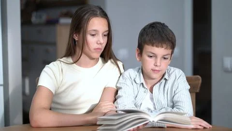 Siblings share a quiet reading session indoors. Focused girl and smiling bo.. Stock Photos