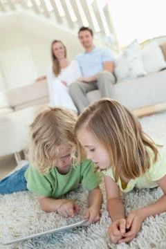 Siblings using tablet on the carpet with parents behind them Stock Photos