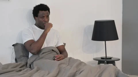 Sick African Man Coughing while Sitting in Bed Stockbeeldmateriaal 299754264