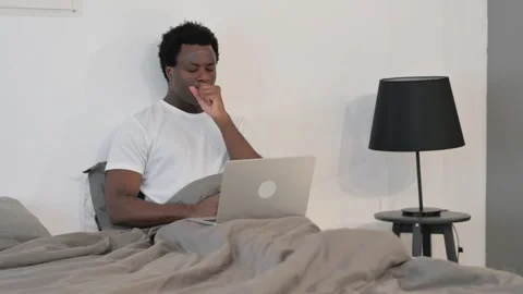 Sick African Man Coughing while Working on Laptop while Sitting in Bed Stockbeeldmateriaal 299755009