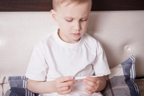 Sick boy sitting in bed and looking at digital thermometer. Ill boy is measur Stock Photos