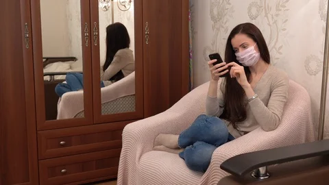 Sick girl in face mask using smartphone, sitting relaxed on armchair. Pandemic Stock Footage 128548444