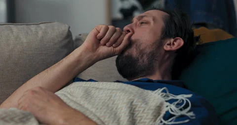 Sick man with beard is lying on sofa and sleeping on blue pillow. Brunette 库存影片 218326115