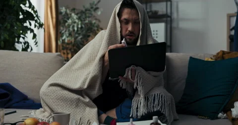 Sick man with beard is sitting on sofa, holding laptop and having video chat 스톡 동영상 218332042
