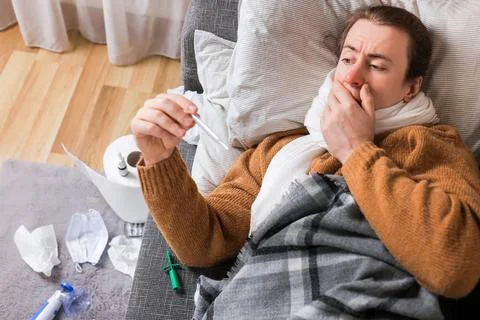Sick man with flu checking thermometer while lying on couch surrounded by t.. Stock Photos