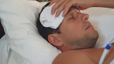 A sick man lies in a bed with a thermometer and a towel on his head, from a Stock Footage 90421790