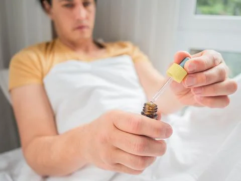 Sick man is lying in bed with liquid medicine. Man is measuring drops. Feelin Stock Photos