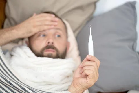 Sick man lying on sofa checking his temperature at home in the living room. M Stock Photos