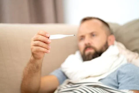 Sick man lying on sofa checking his temperature at home in the living room. Stock Photos