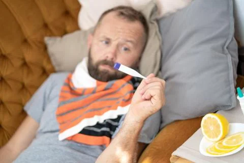 Sick man lying on sofa checking his temperature at home in the living room. M Stock Photos