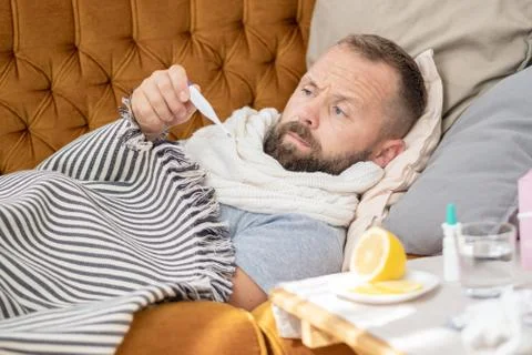 Sick man lying on sofa checking his temperature at home in the living room. M Stock Photos