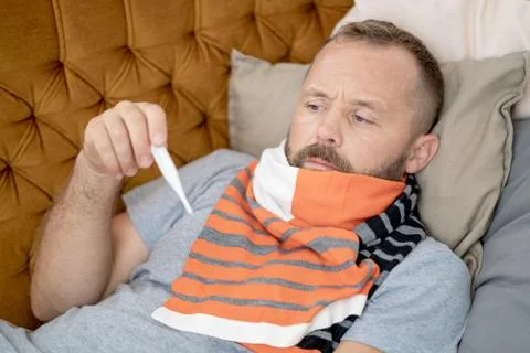 Sick man lying on sofa checking his temperature at home in the living room. M Stock Photos