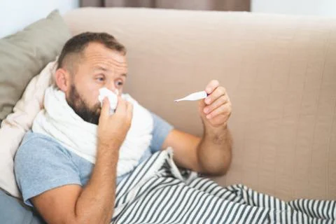 Sick man lying on sofa checking his temperature at home in the living room. M Stock Photos