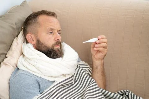 Sick man lying on sofa checking his temperature at home in the living room. M Stock Photos