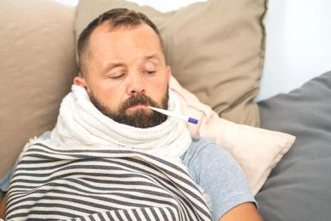 Sick man lying on sofa checking his temperature at home in the living room. M Stock Photos