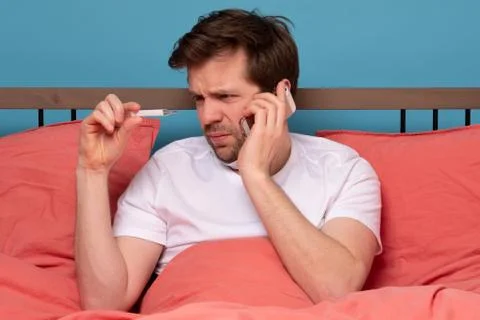 Sick man lying on sofa checking his temperature at home and calling to his do Stock Photos