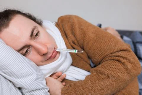 Sick man in scarf lying on sofa checking high fever temperature with thermo.. Stock Photos