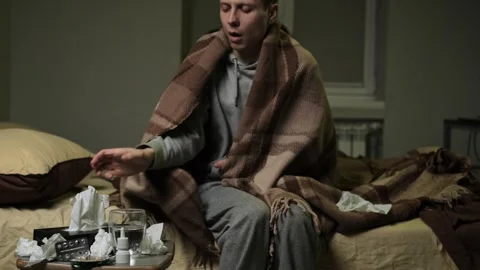 Sick man sits on a bed under blanket, sneezing into a tissue Stock Footage 297863083