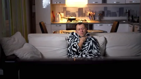 A sick man sits on the couch in the evening and watches TV, camera movement Stock Footage 131889339