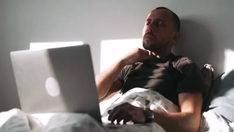 A sick man takes his temperature and works on a laptop while sitting in bed The Stock Footage 154342647