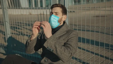 Sick man wearing Protective Face Mask sitting near fence border sneeze coughs Stock Footage 126617739