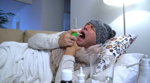 Sick man wearing a warm cap and scarf in a bed coughs and uses spray for throat. Stock Footage 68932573