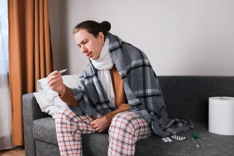 Sick man wrapped in blanket checking thermometer on sofa at home with medic.. Stock Photos