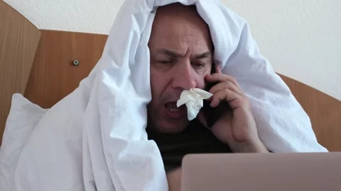 Sick middle aged man sitting on bed with laptop. He has a tissue in his nose Stock Footage 301892944