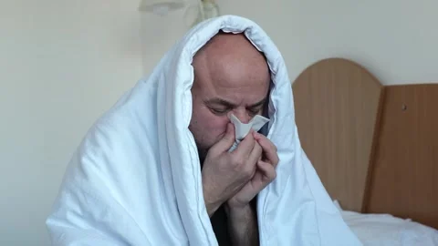 Sick middle aged man sitting on bed in bedroom or in hotel room and blowing nose Stock-Footage 310712334