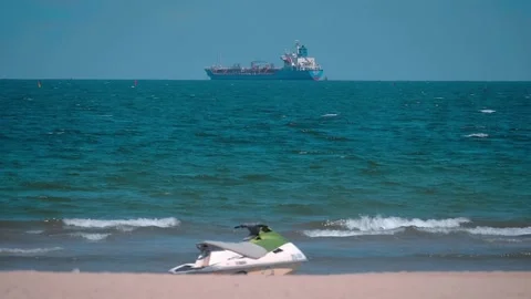 Sick ocean-going cargo ship anchored far of a beach where the jet ski is parked Stock Footage 303172574
