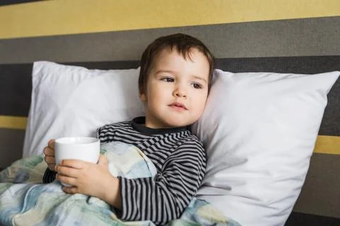 Sick upset curing boy with cup of medicament lying on bed Stock Photos