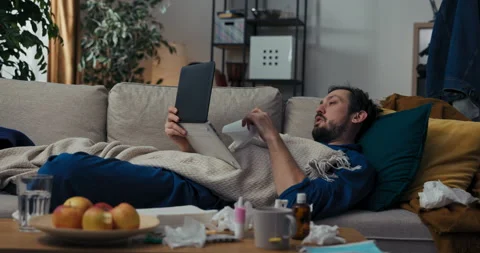 Sick young man with beard and black hair is lying on sofa and covered with Stockbeeldmateriaal 218323336