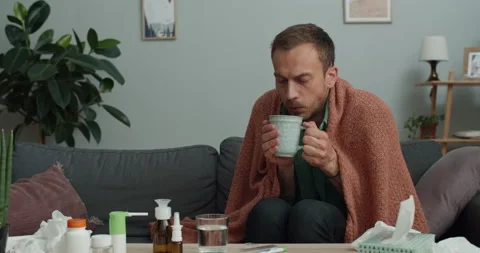 Sick young man blowing on cup with hot tea and drinking while sitting on couch Stock Footage 142634019
