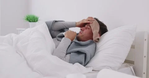Sick young man lying in bed under blanket with wet towel on his head, suffering Stock Footage 287568470