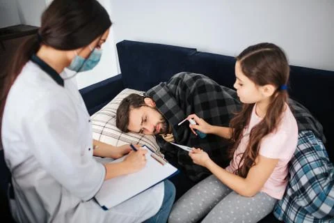 Sick young man lying on bed. His daughter and female doctor sitting besides 库存照片