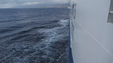 Side of large ferry boat, bow waves on dark blue sea and coastal town on horizon Stock Footage 99003568