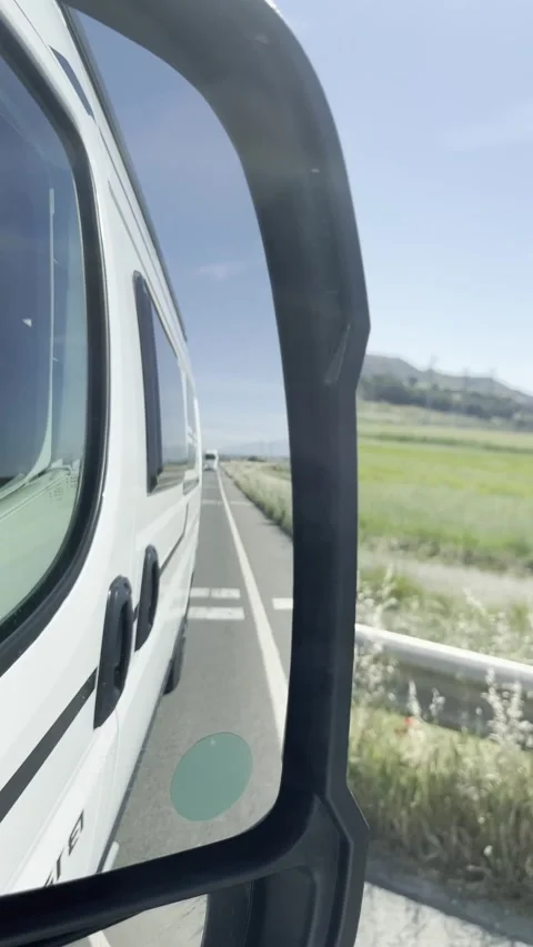 Side mirror reflection of a van driving along a road lined with greenery under a Stock Footage 346326730