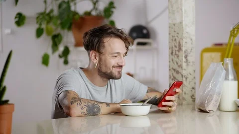 Side portrait of cute man eating breakfast and using red gadget online in social Stock Footage 108798473